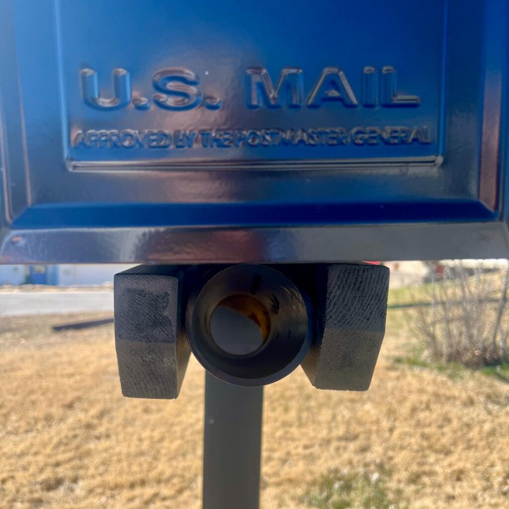 Close-up of a blue U.S. Mail collection box with its chute covered by two metal brackets. The engraved words “U.S. MAIL APPROVED BY THE POSTMASTER GENERAL” are visible on the box’s front. Grass and a road are in the background.