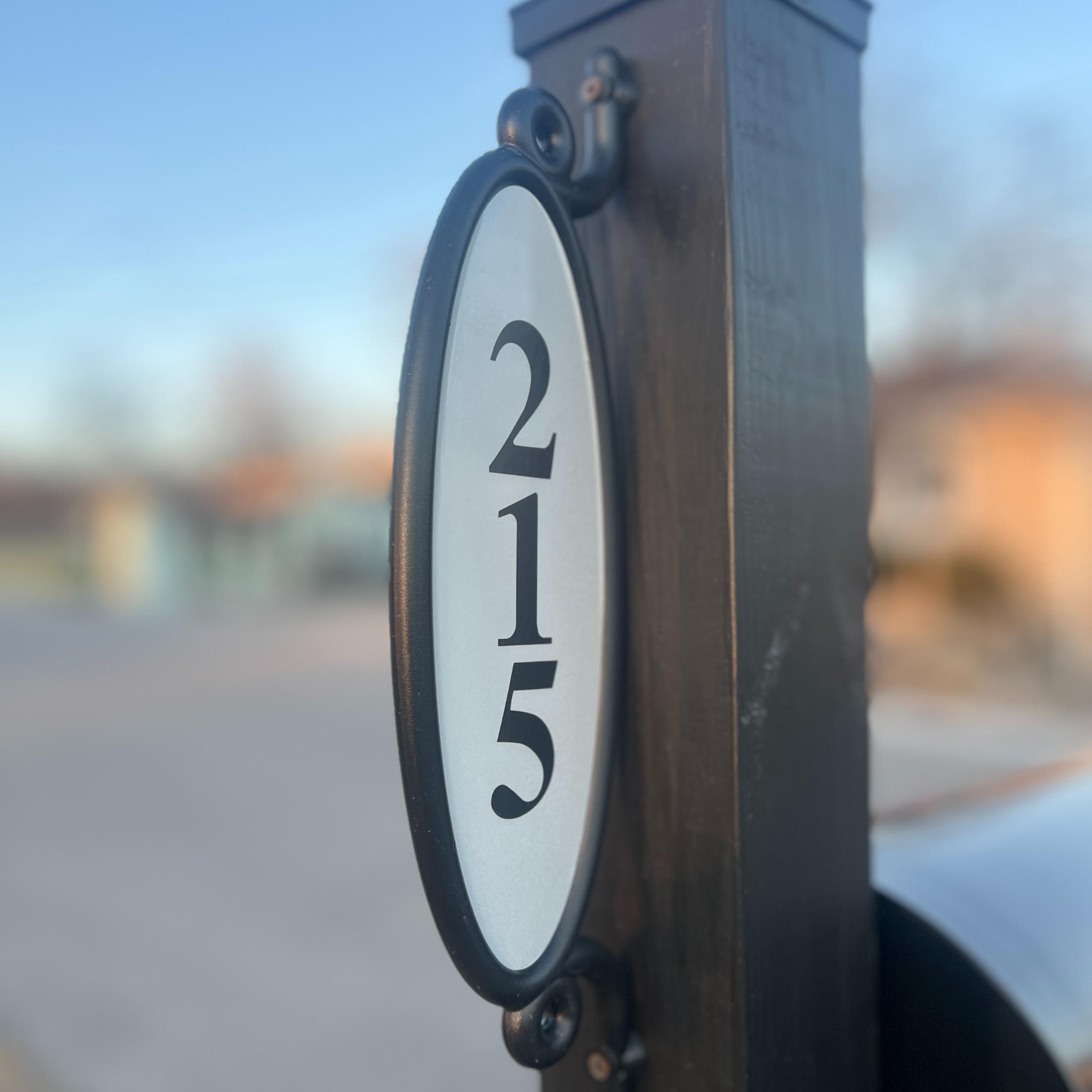 A close-up of a black and white oval house number sign displaying the numbers "215" mounted on a dark post, with a blurred suburban street background.