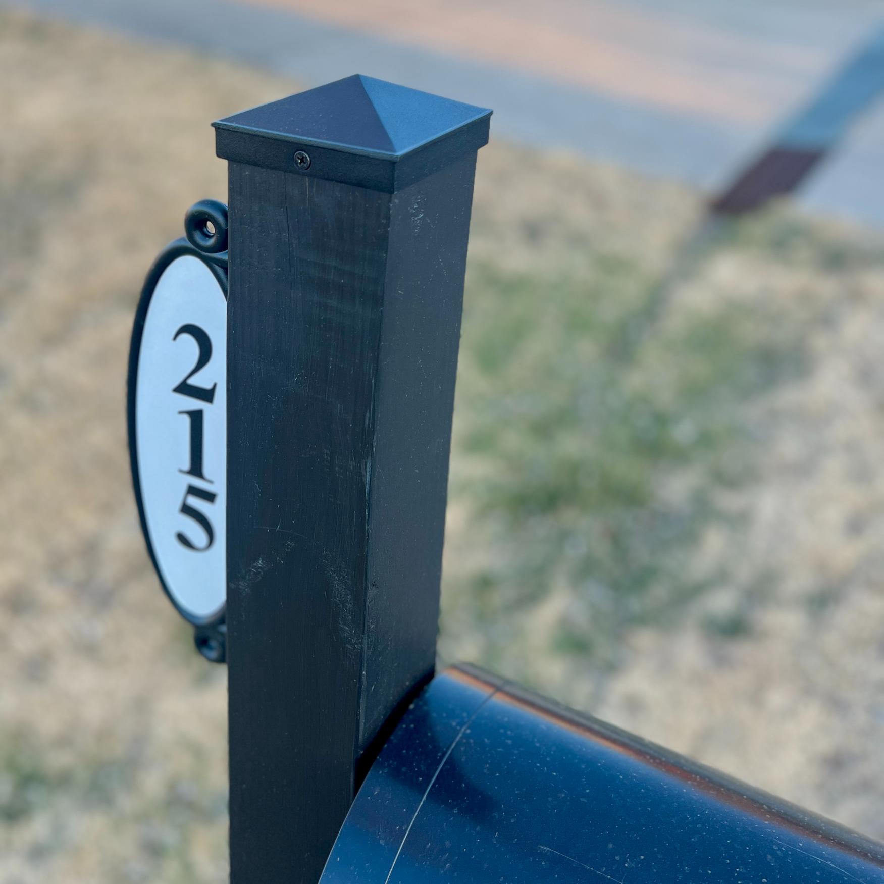A black mailbox post with a white oval address plaque displaying the numbers "215" in black, set against a blurred background of grass and pavement.