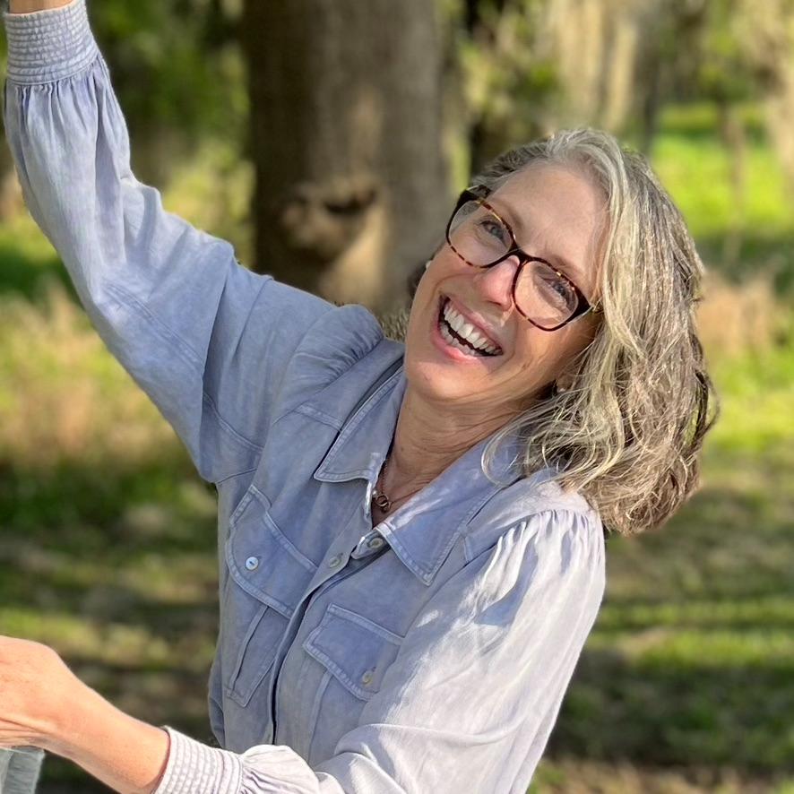 A smiling woman with gray hair and glasses wearing a light blue shirt stands outdoors in sunlight, with a blurred background of trees and grass.