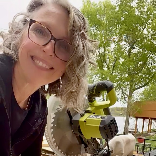A smiling woman with curly gray hair and glasses stands next to a green Ryobi miter saw outdoors, with trees and a lakefront cabin visible in the background.