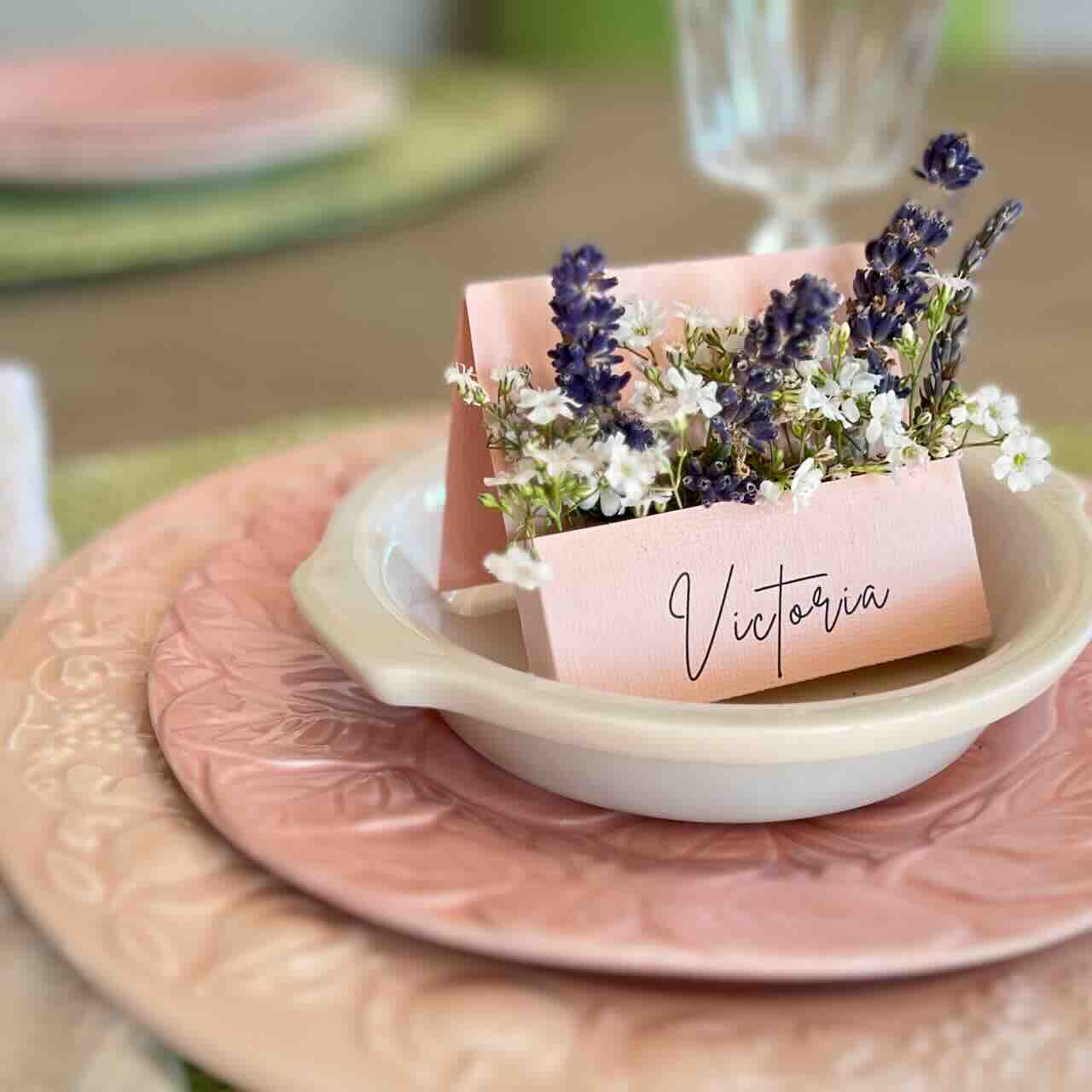 A place setting with pastel pink plates, a white bowl, and a folded name card reading “Victoria,” decorated with small white flowers and sprigs of lavender. The table has a soft, elegant, spring-like appearance.