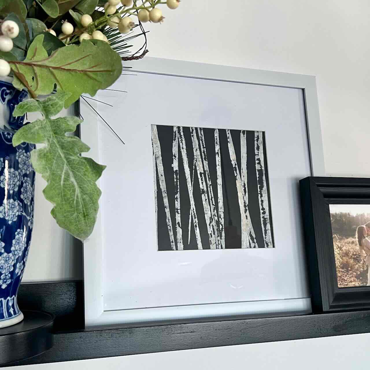 A framed black and white artwork of birch trees sits on a black shelf next to a blue and white vase with green leaves and berries, and a framed photograph.