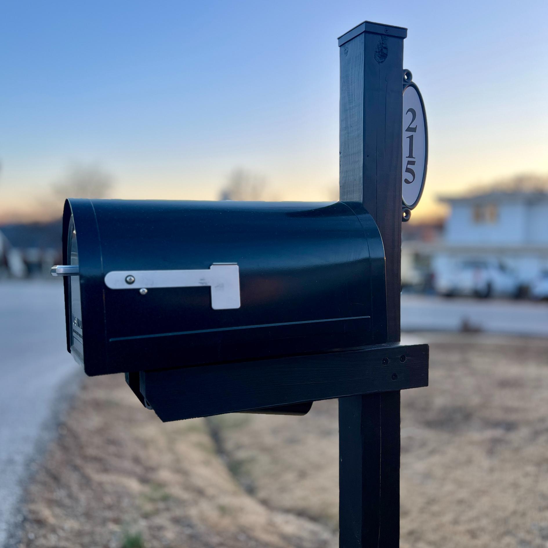 A black mailbox with a white flag stands on a post labeled "215" in a suburban neighborhood at sunset, with blurred houses and cars in the background.