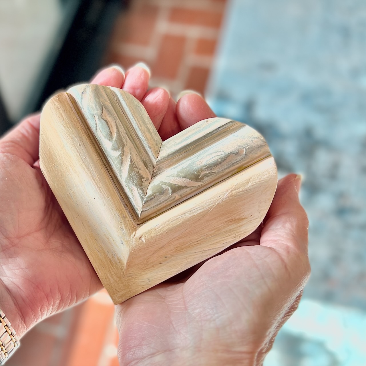 A pair of hands gently holds a carved wooden heart with a marbled pattern on one side, against a softly blurred background of tile and blue surfaces.