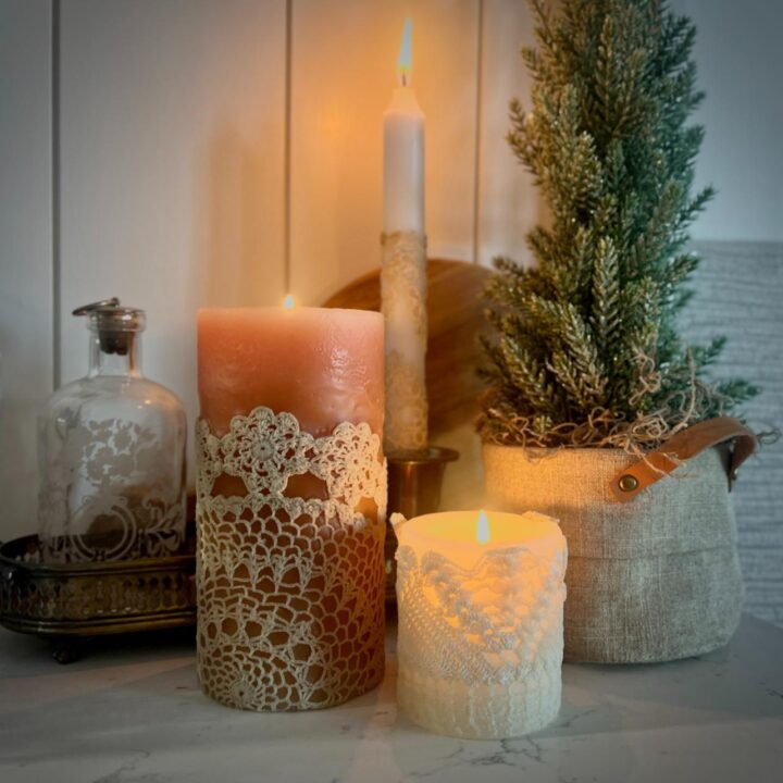 Three DIY lace candles burn beside a small potted pine tree and a glass bottle on a marble surface, creating a cozy, rustic setting with soft, warm lighting.