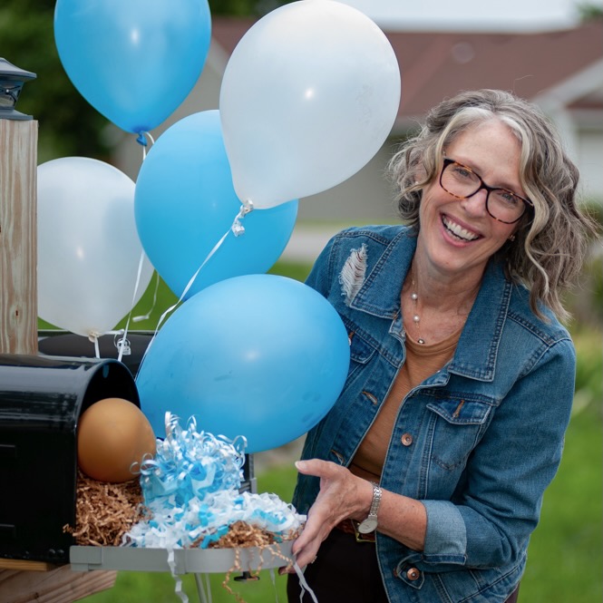 A smiling woman with gray hair and glasses, wearing a denim jacket, stands by an open mailbox decorated with blue and white balloons and festive shredded paper. She gestures toward the mailbox contents outdoors.