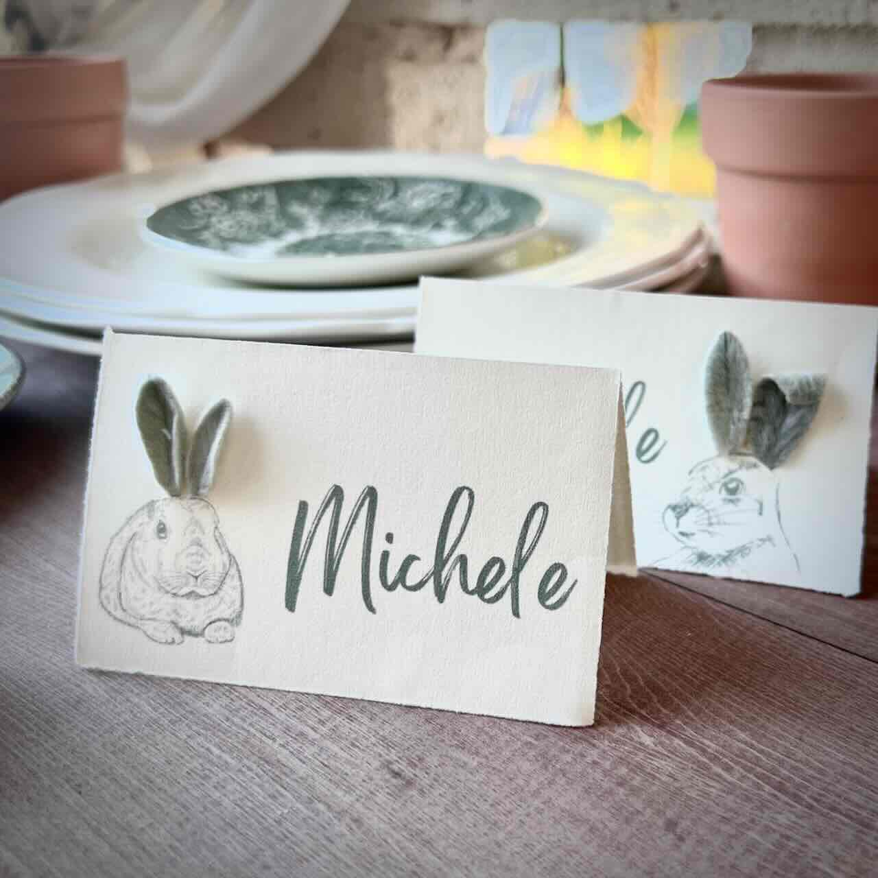 Two place cards with bunny drawings and gray felt ears stand on a wooden table. One card reads "Michele." Plates and terracotta pots are in the background.