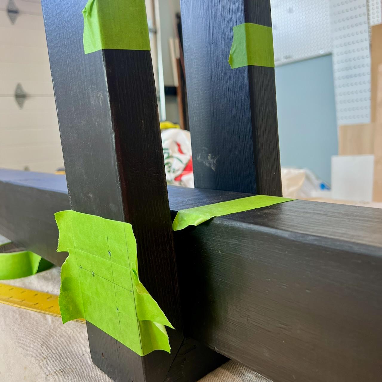 Close-up of a woodwork project with dark wooden beams joined at a right angle, secured with green painter’s tape. Workshop materials and tools are visible in the blurred background.