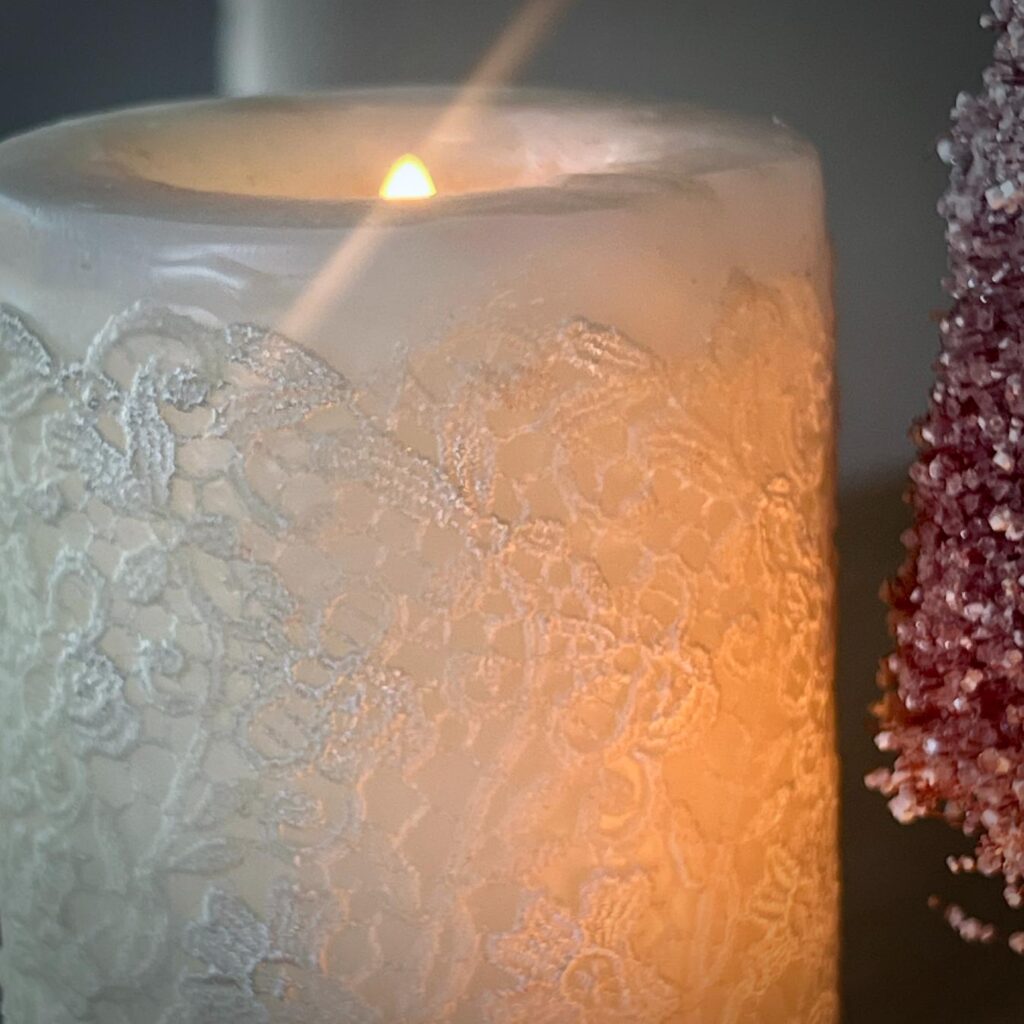 A white DIY lace candle is lit, casting a warm glow. A red, glittery decorative object is partially visible on the right side of the image.