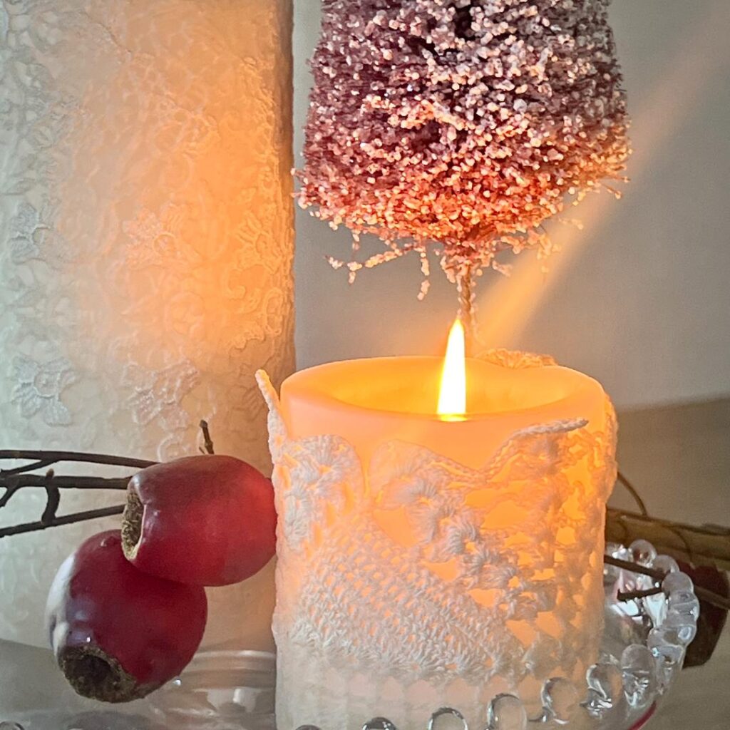 A lit DIY lace candle sits next to two red berries and a branch. Above the candle is a frosted, purple bottle-brush tree. Soft, warm lighting creates a cozy atmosphere.