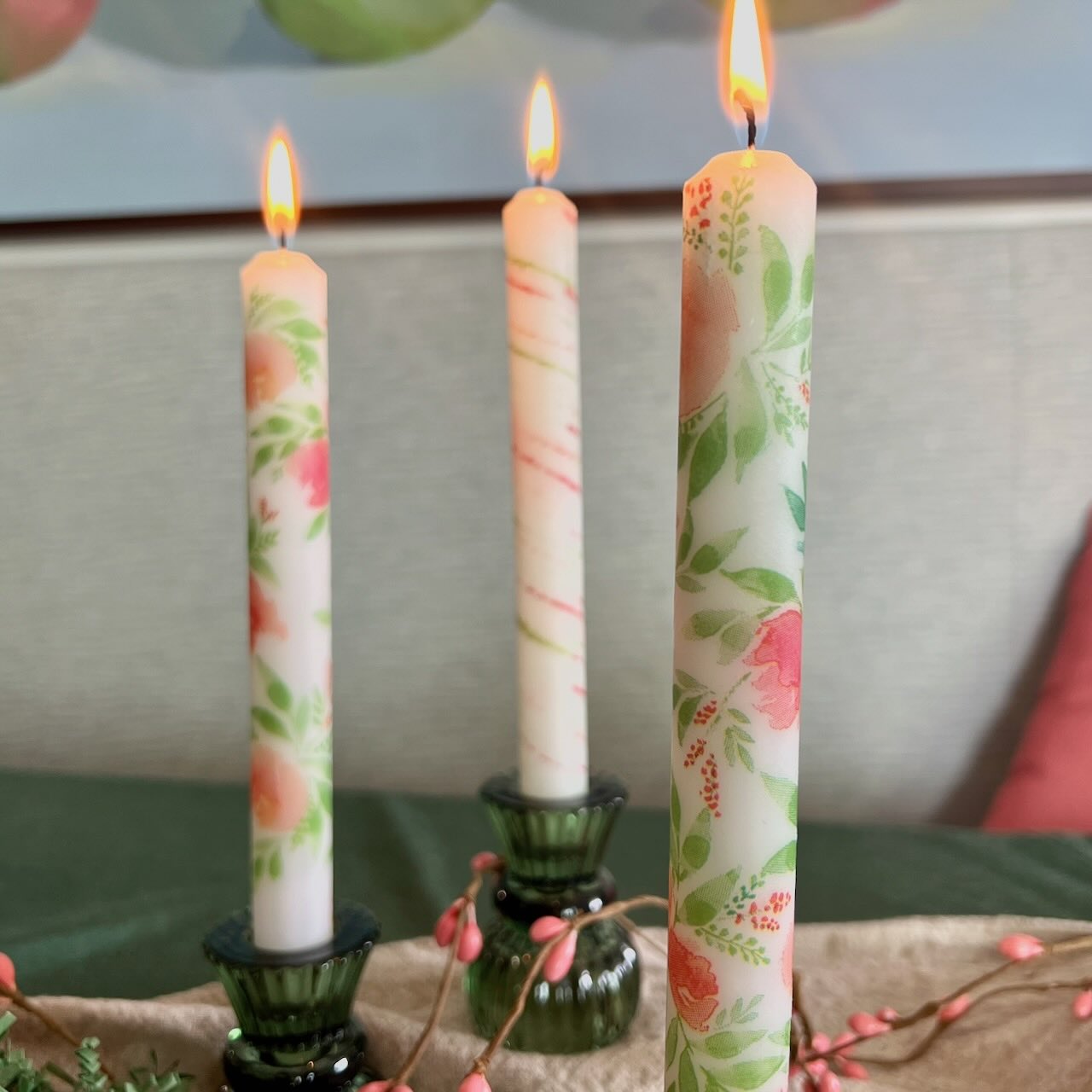Three lit candles with floral and striped designs are placed in green glass holders on a table, with a soft-focus background featuring a painting and a red pillow. Pink decorative berries lie on the table.