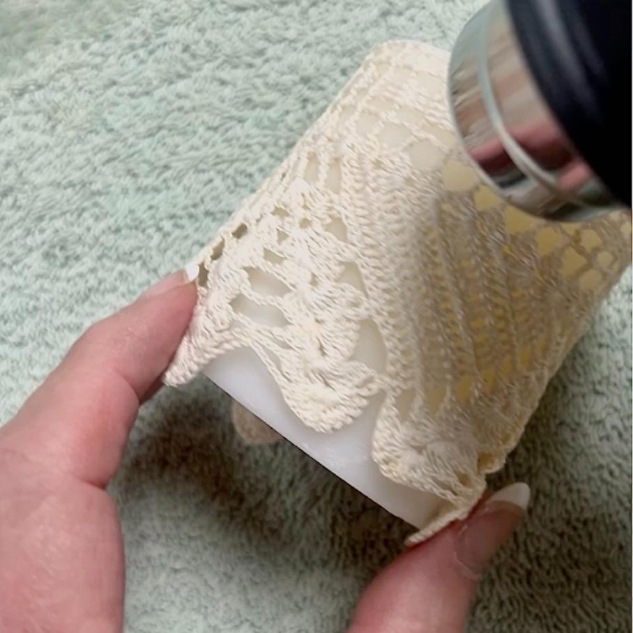 A hand holds a white cylindrical object wrapped in beige crochet lace while a heat tool is applied to the surface, creating beautiful DIY lace candles on a light green textured background.