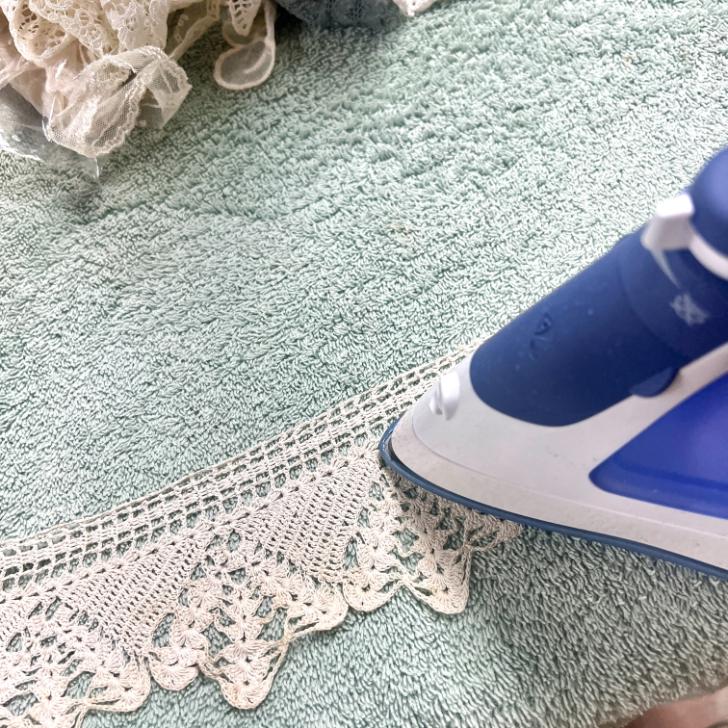 A close-up of a blue and white iron pressing a piece of lace on a light green towel, with more lace fabric in the background—perfect for prepping materials for DIY lace candles.