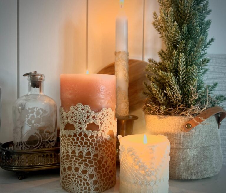 A cozy scene with DIY lace candles glowing softly, a small potted evergreen tree, a decorative glass bottle on a tray, and a wooden board set against a white paneled background.