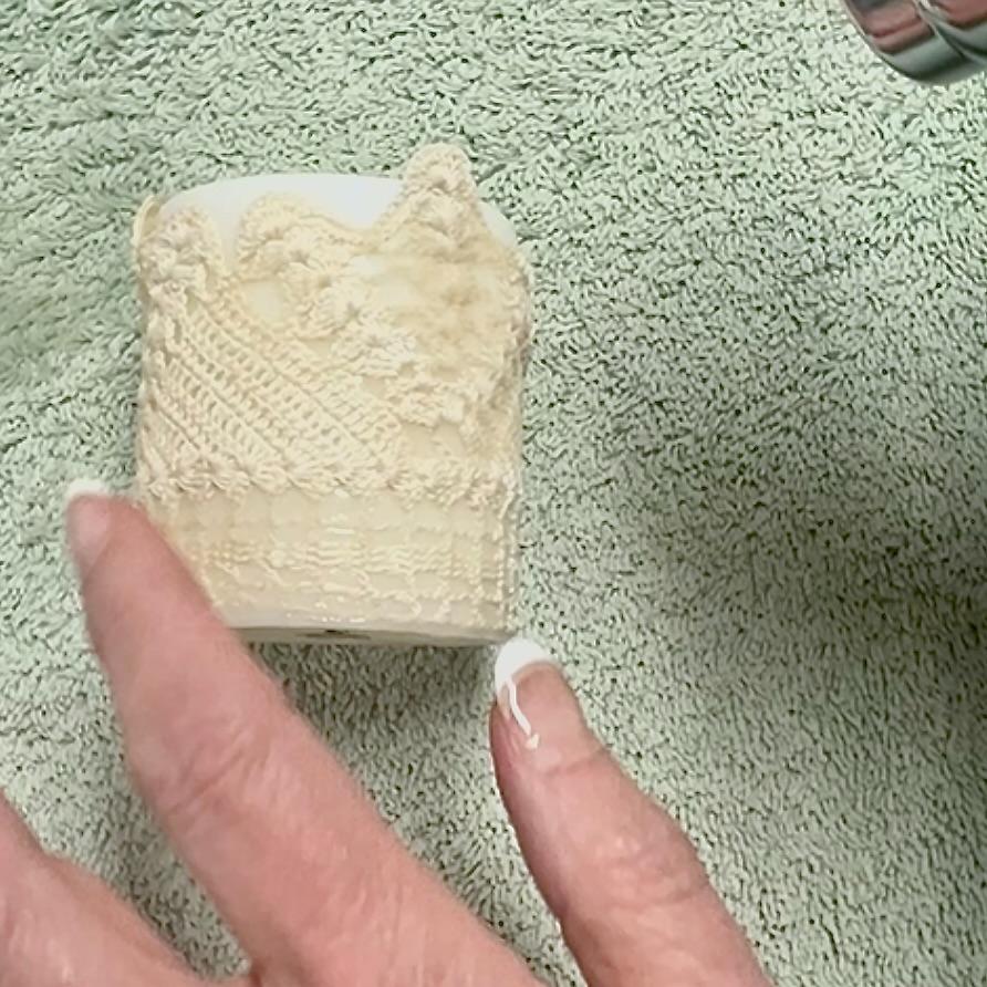 A hand with a French manicure holds a white cup, decorated like elegant DIY lace candles, positioned on a light green textured towel.