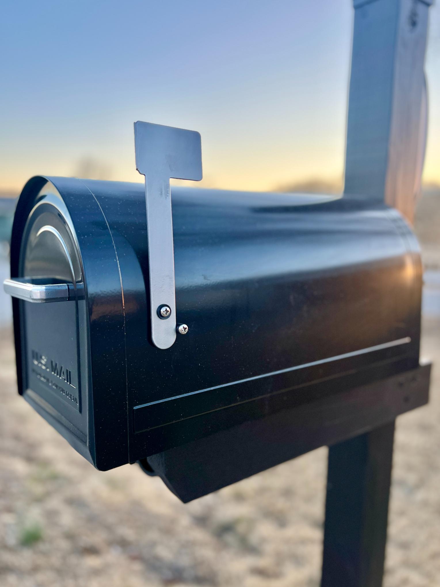 A black metal mailbox with the flag up is mounted on a post outdoors. The background shows a blurry landscape at sunset.