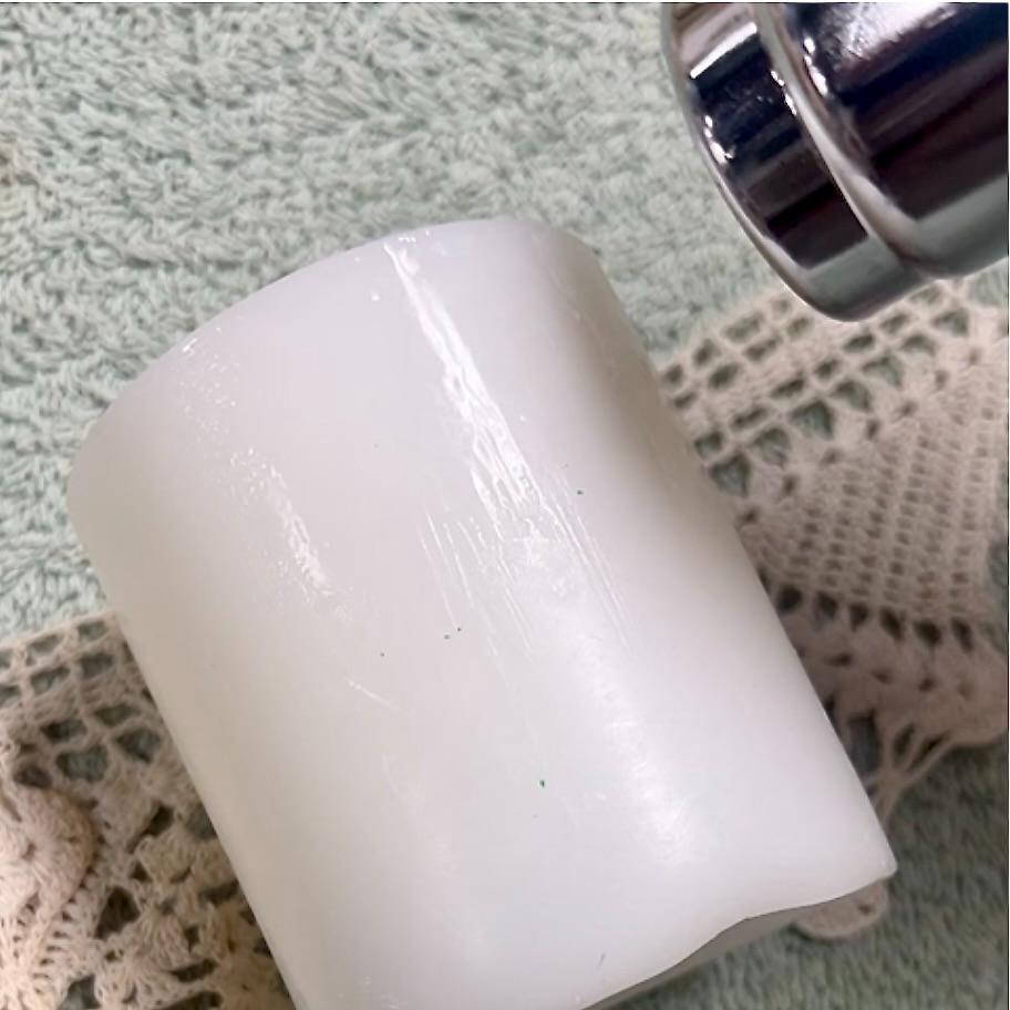 A silver hair dryer blows warm air onto a large white wax cylinder, perfect for DIY lace candles, resting on a textured, light-colored surface layered with lace and knit fabrics underneath.