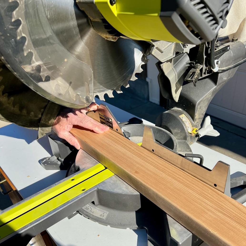 A person uses a miter saw to cut a piece of wood. The wood is held firmly in place on the saw table, and sunlight brightens the workspace.