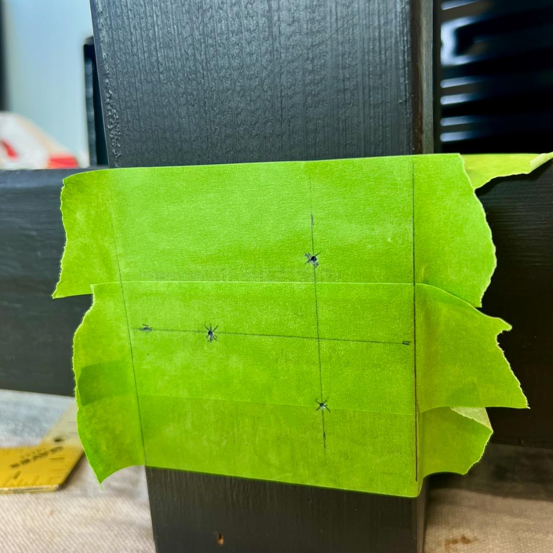 A piece of green painter’s tape is attached to a dark wooden surface, marked with intersecting pencil lines and three drilled cross points, likely for precise drilling or hardware placement.