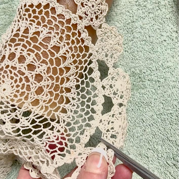 A hand holding scissors is cutting the edge of an intricate cream-colored crocheted doily, which is laid on a light green textured fabric—perfect for DIY lace candles.