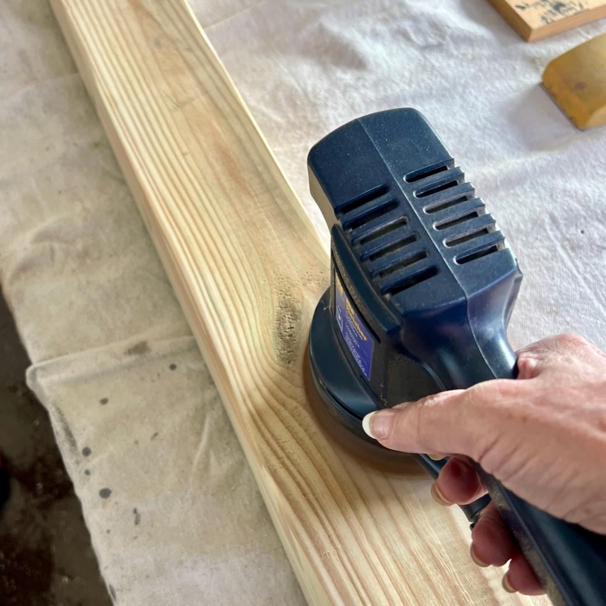 A hand uses a blue electric sander on a long wooden plank, smoothing the surface. The background shows a white cloth underneath the wood.