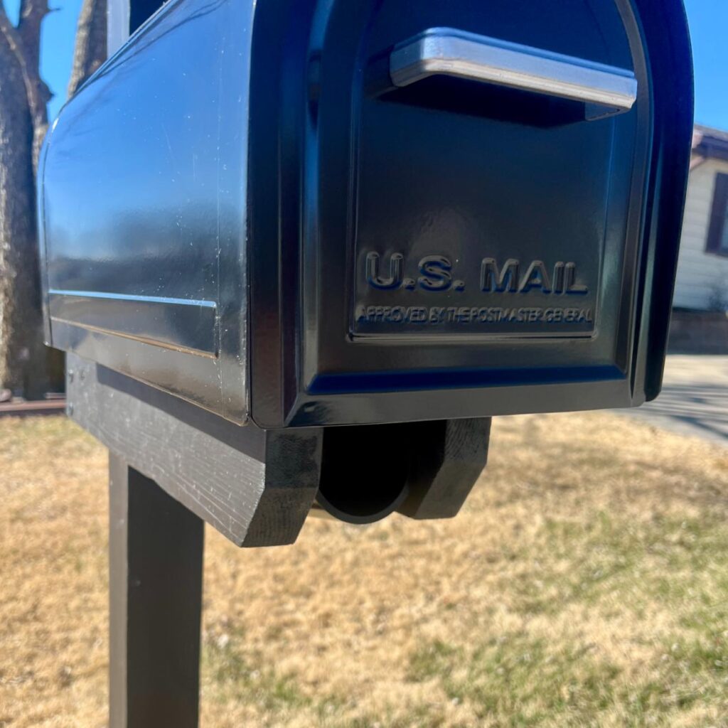 Close-up of a black mailbox with “U.S. MAIL” embossed on the front, mounted on a black post, standing in a yard with dry grass and a house visible in the background.
