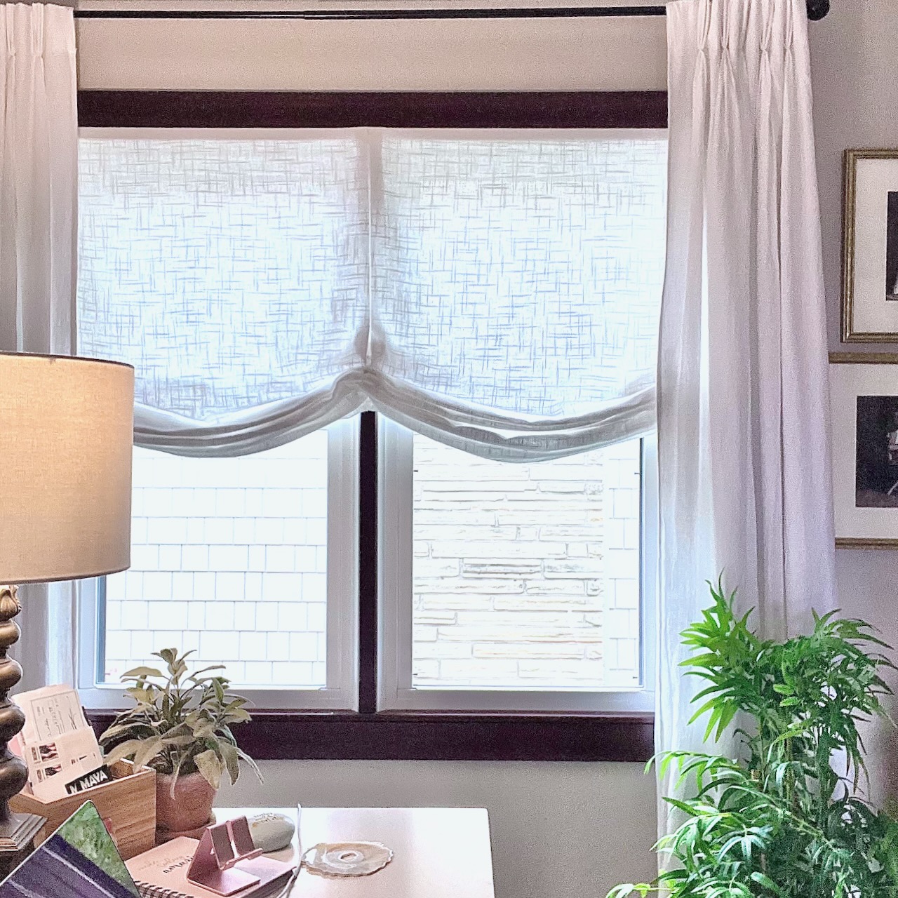 A bright room with a window covered by sheer white curtains, a desk with papers and a lamp on the left, potted plants on the desk and floor, and framed artwork partially visible on the right wall.