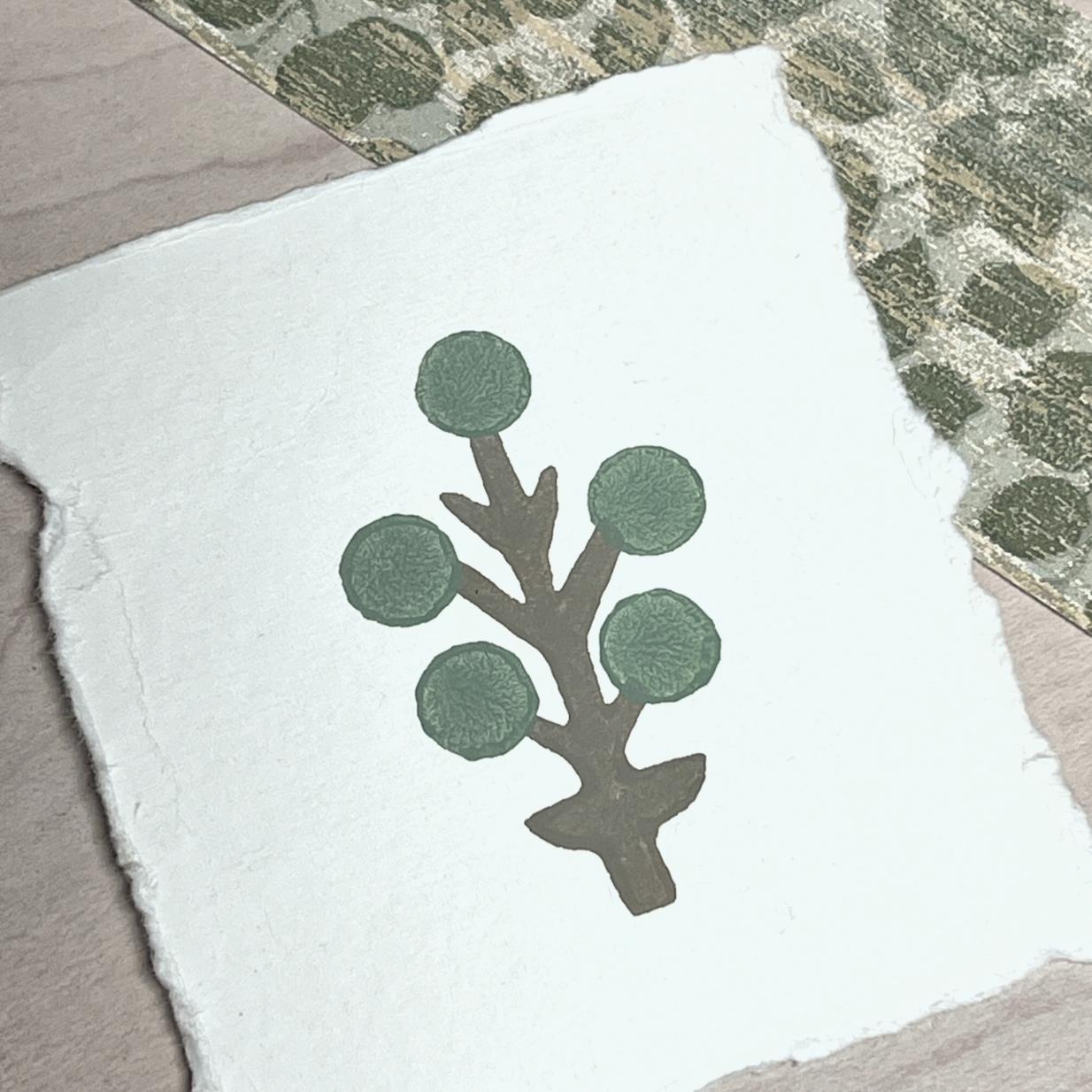 A piece of textured white paper with a simple illustration of a brown branch and five green circular leaves, placed on a light wood surface near patterned green and beige fabric.