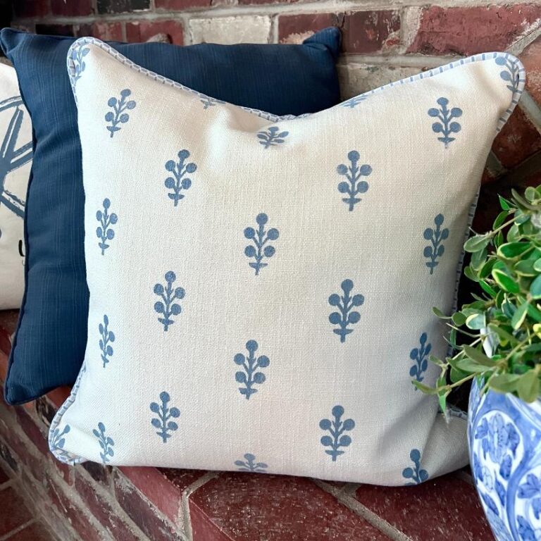 A decorative pillow with a light beige background and blue leaf-like patterns sits on a brick bench next to dark blue pillows and a blue and white ceramic pot with a green plant.