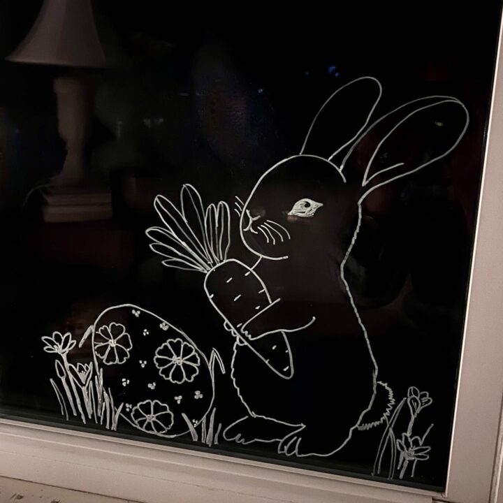 A white line drawing on glass shows a rabbit holding a large carrot next to a decorated Easter egg and some grass and flowers, with a lamp faintly visible in the background.