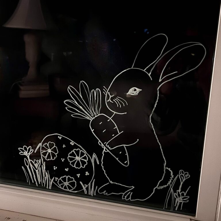 A white line drawing on glass shows a rabbit holding a large carrot next to a decorated Easter egg and some grass and flowers, with a lamp faintly visible in the background.