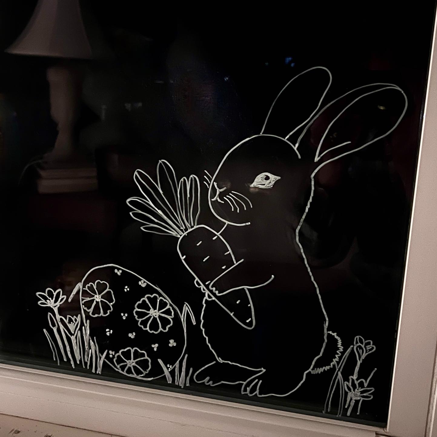 A white line drawing on glass shows a rabbit holding a large carrot next to a decorated Easter egg and some grass and flowers, with a lamp faintly visible in the background.