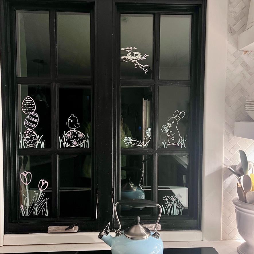 A black-framed kitchen window decorated with white drawings of Easter eggs, a chick, a bunny, a bird’s nest, tulips, and grass. A blue kettle sits on the stove below the window.