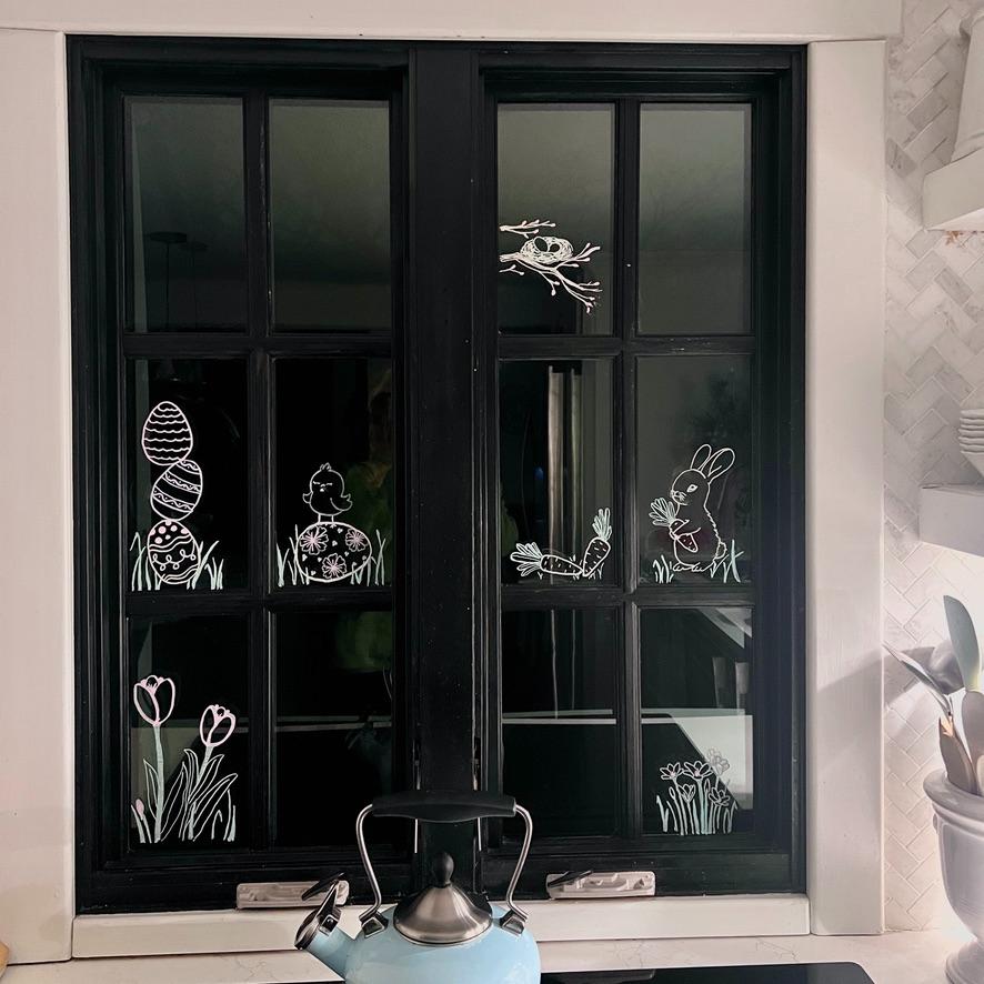 A kitchen window decorated with white drawings of Easter eggs, a chick, a rabbit, tulips, grass, and a bird in a nest. A blue kettle sits on the stove below the window.