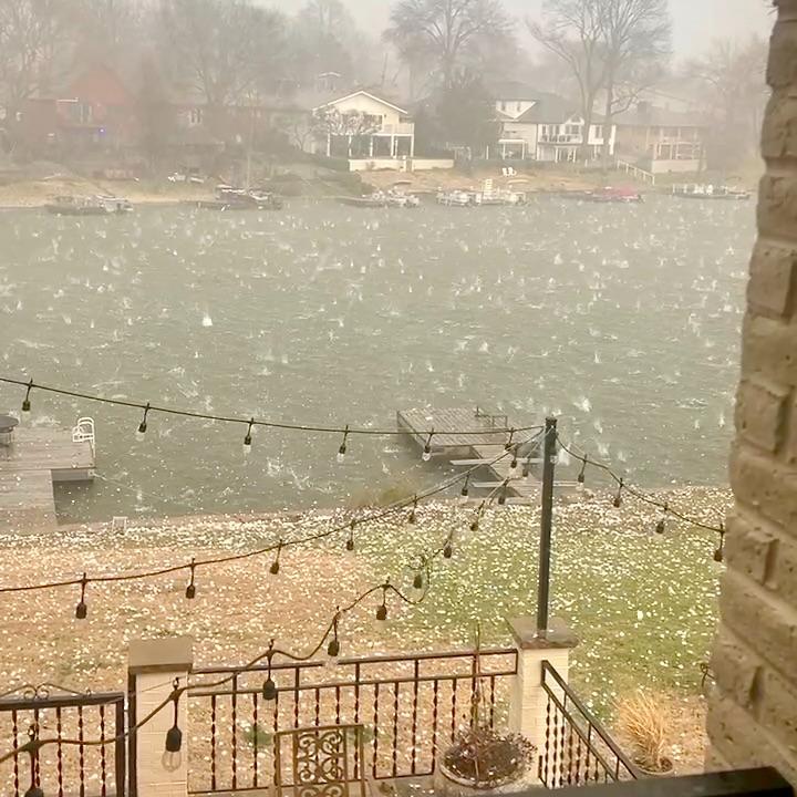 Large hailstones fall heavily on a lakeside neighborhood, bouncing off the grass and dock, with string lights hanging in the foreground and houses and trees visible across the water under a cloudy sky.