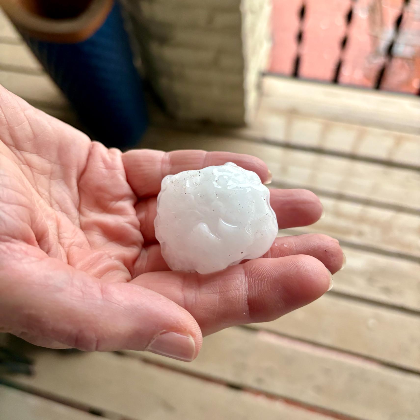 A person holding a large, irregularly shaped piece of hail in their palm, standing on a wooden deck with a brick wall and railing in the background.