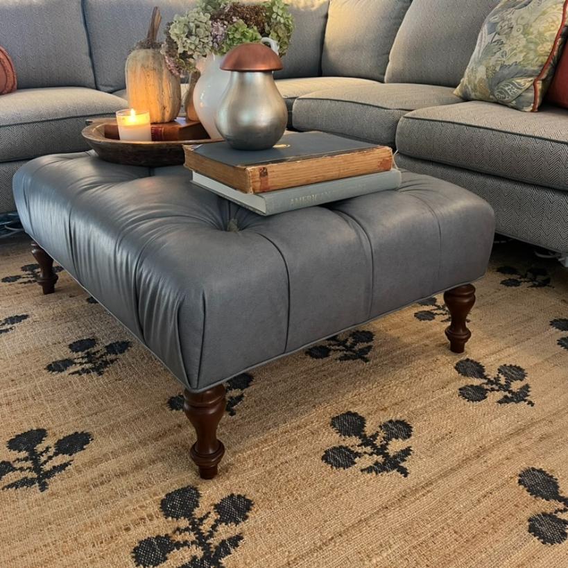 A gray tufted ottoman with wooden legs is used as a coffee table, holding stacked books, a tray with a candle, vase, and decorative items. It sits on a tan rug with black floral patterns in front of a gray sectional sofa.