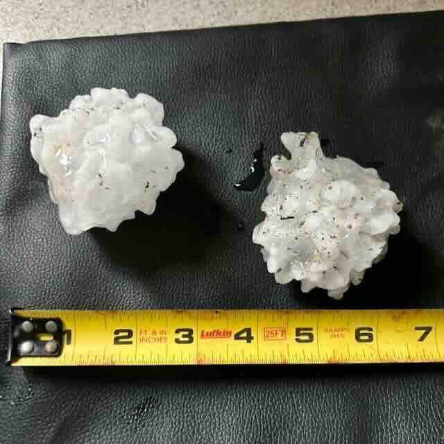 Two large, irregularly shaped hailstones rest on a black surface next to a yellow measuring tape. The hailstones are each about 3 inches wide.
