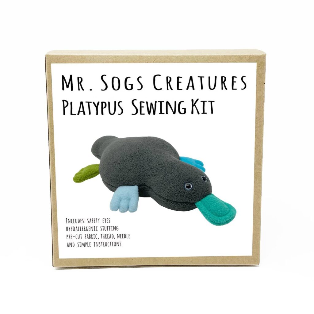 A cardboard box labeled “Mr. Sogs Creatures Platypus Sewing Kit” features a photo of a stuffed platypus. Text notes the kit includes safety eyes, thread, needle, pre-cut fabric, stuffing, and instructions.