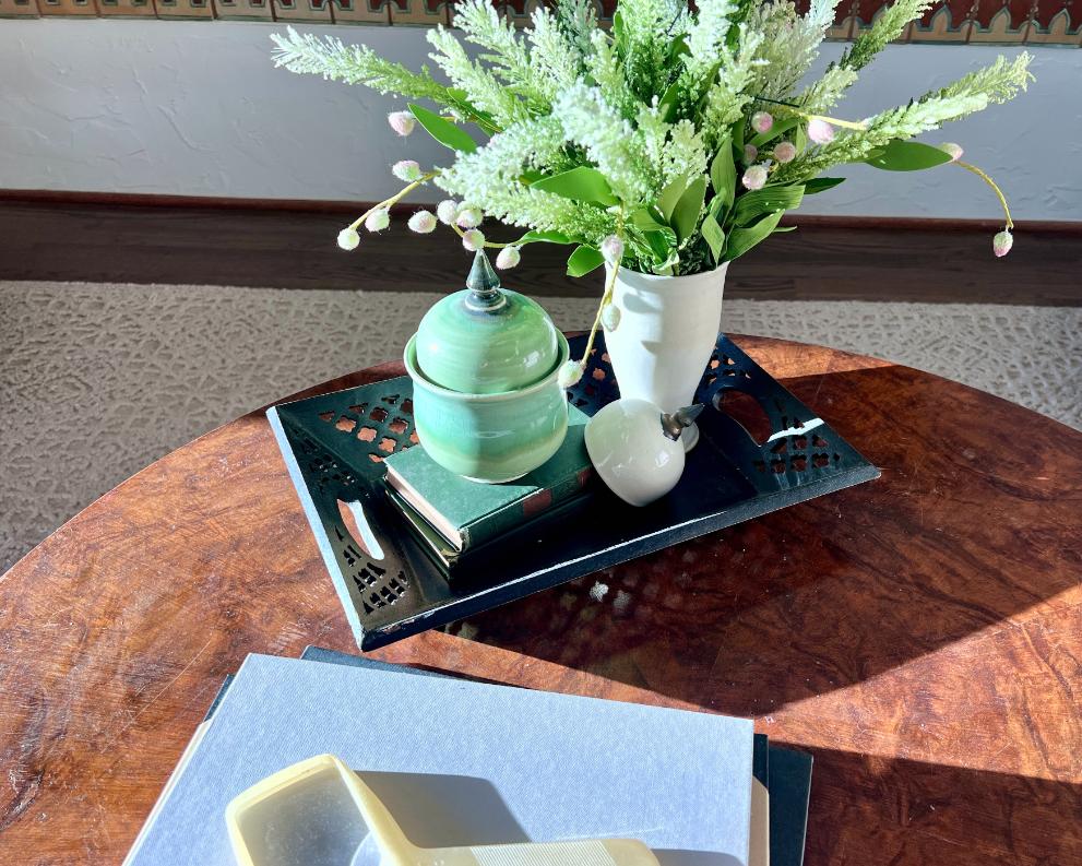 A green glass jar, a white vase with faux white flowers, and a small white apple sit on a black tray atop a round wooden table. Documents and a magnifying glass lie on the table in sunlight.