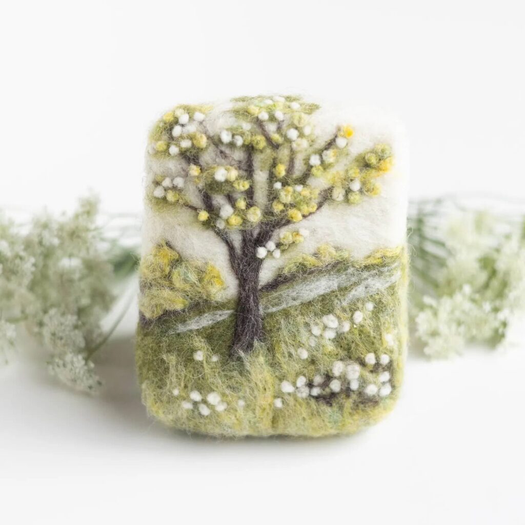 A rectangular felted soap bar decorated with a needle-felted spring landscape, featuring a blossoming tree with yellow and white flowers, green grass, and delicate floral details. Pale flowers are blurred in the background.