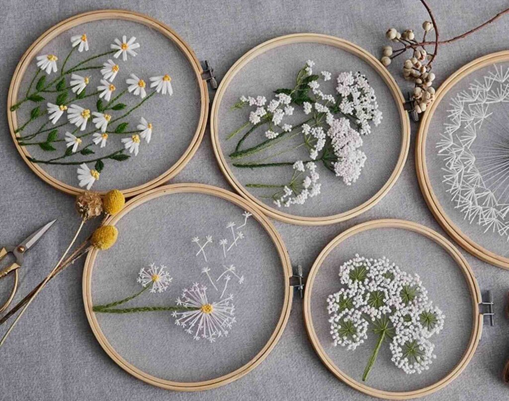 Five embroidery hoops with delicate floral designs stitched on sheer fabric are arranged on a grey surface. Nearby are a pair of scissors, two yellow billy balls, and some dried berries.