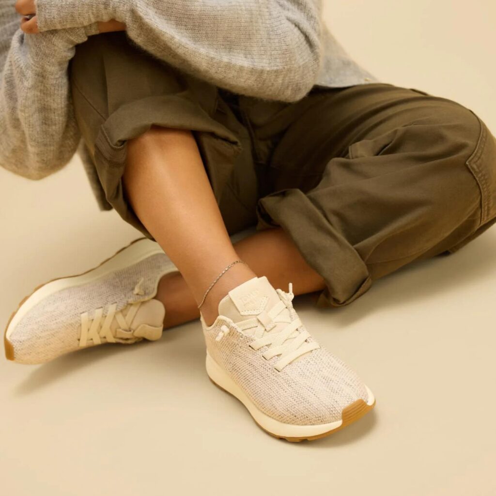 A person wearing olive green pants, a gray sweater, and beige knit sneakers with gum soles sits on the floor with legs crossed. Only their lower body is visible against a neutral background.