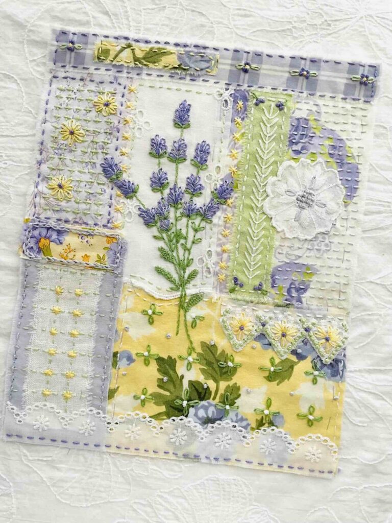 A patchwork textile art piece featuring hand-stitched lavender, daisies, and leaves in purple, yellow, and green fabrics, with lace and embroidered details on a white textured background.