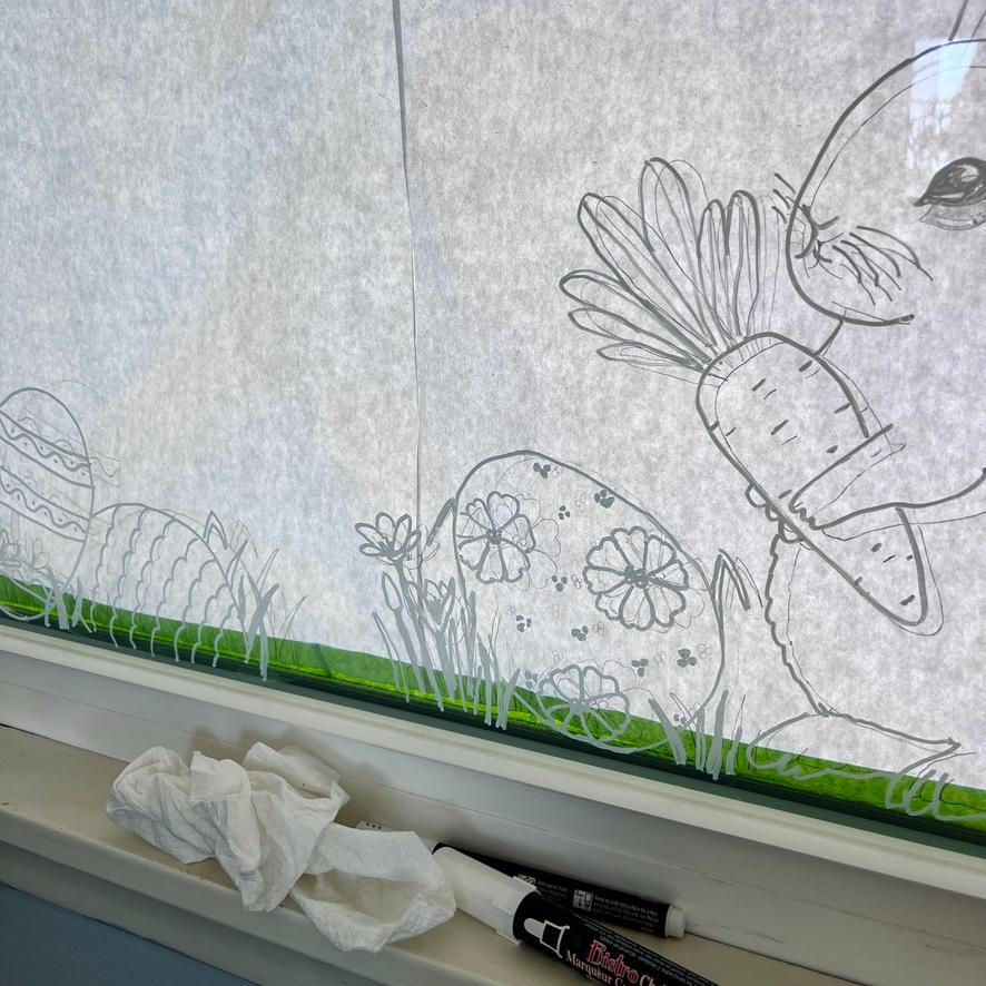 A window decorated with white marker drawings of a rabbit holding a carrot, decorated Easter eggs, and grass. Tissues and marker pens are on the windowsill below.