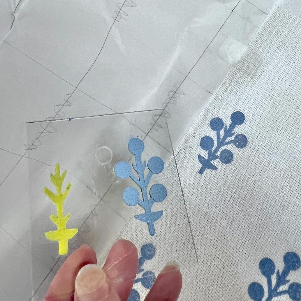 A hand holds a clear stencil with a plant design. The stencil has yellow and blue paint stains. The same blue plant pattern is stamped repeatedly on a piece of white fabric beneath.