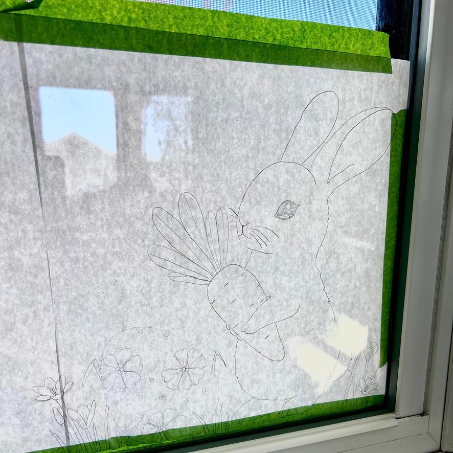 A hand-drawn outline of a rabbit holding a large carrot, taped to a window with green tape. Flowers and grass are sketched around the rabbit, and sunlight shines through the paper.