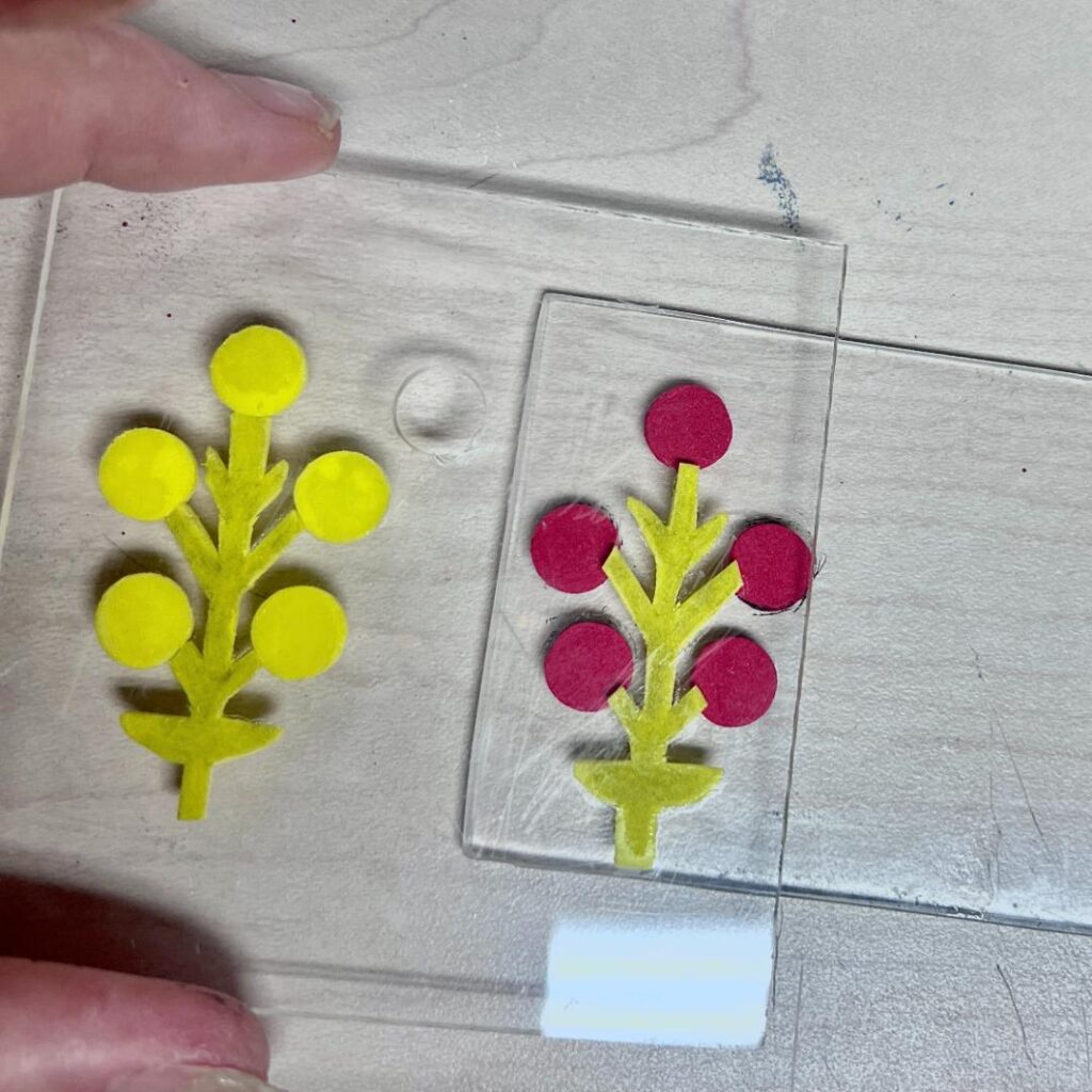 A hand holds a clear panel with a yellow felt flower design, next to a similar red flower design on another clear panel, both on a light wooden surface.