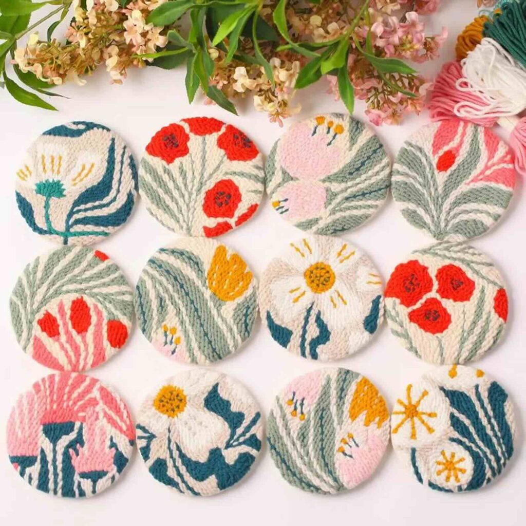 Circular coasters with colorful embroidered floral designs are arranged in rows beneath green leafy branches and flowers. Balls of pink and white yarn are in the upper right corner.
